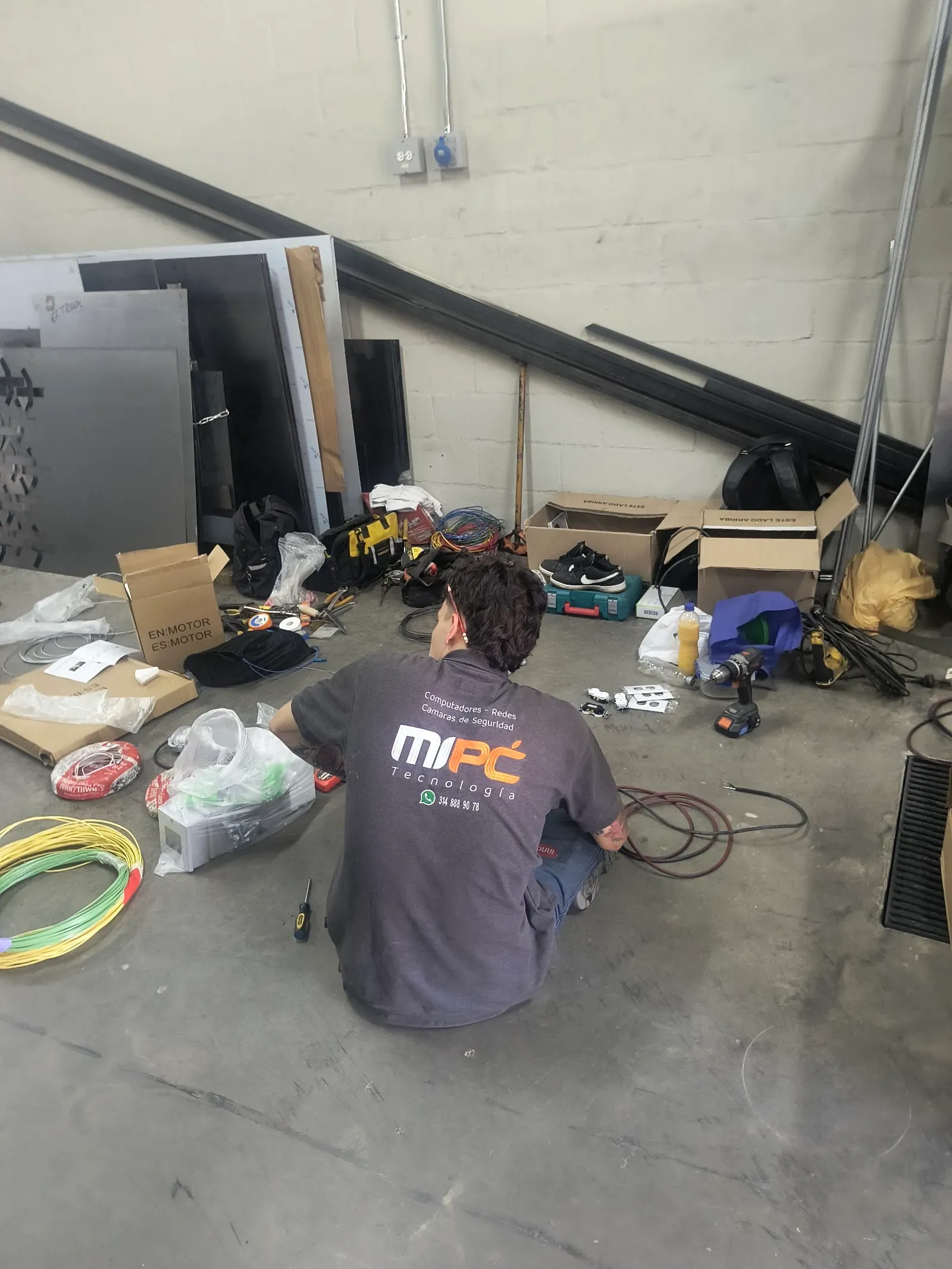 Técnico de MiPC Tecnología prepara el cableado eléctrico sentado en el piso de la bodega en obra, con rollos de cable de fase y de tierra amarillo y verde, tomas sin instalar y herramienta alrededor