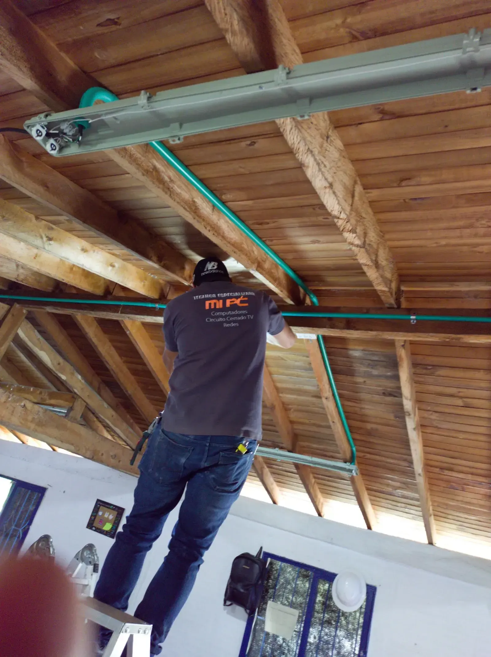 Técnico especializado de MiPC Tecnología tiende canalización verde y una luminaria sobre la estructura de vigas de madera del techo de la edificación