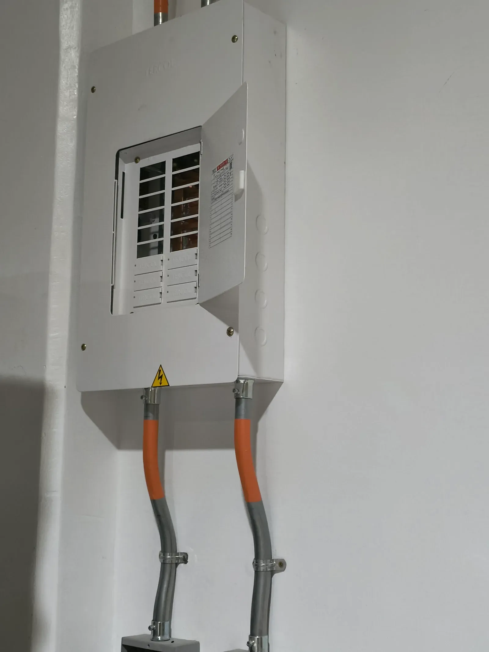 Tablero eléctrico de distribución terminado, con la tapa de los breakers abierta y dos acometidas en tubería metálica sujetas a la pared con abrazaderas