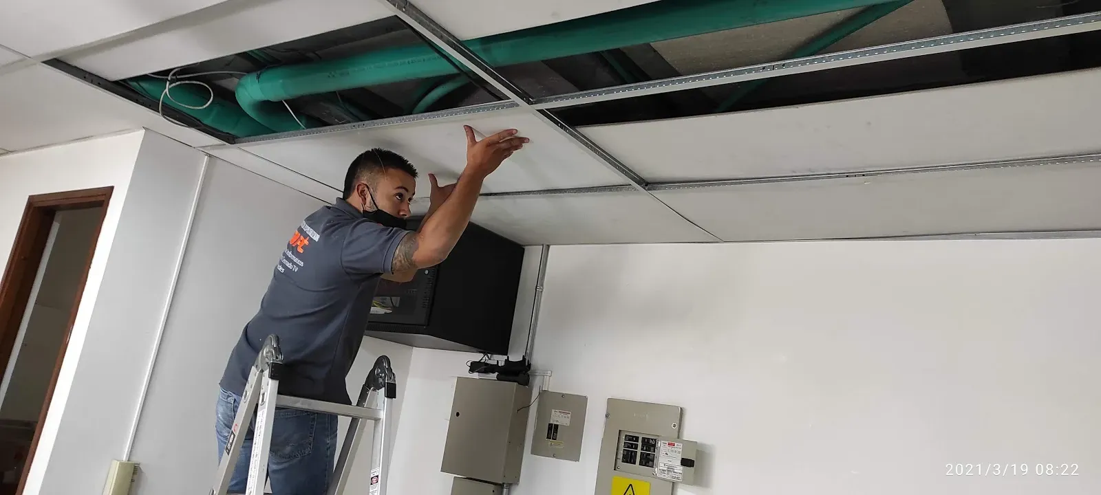 Técnico de MiPC Tecnología subido en una escalera coloca una placa de cielo raso sobre la canalización recién tendida, junto a los tableros eléctricos del piso