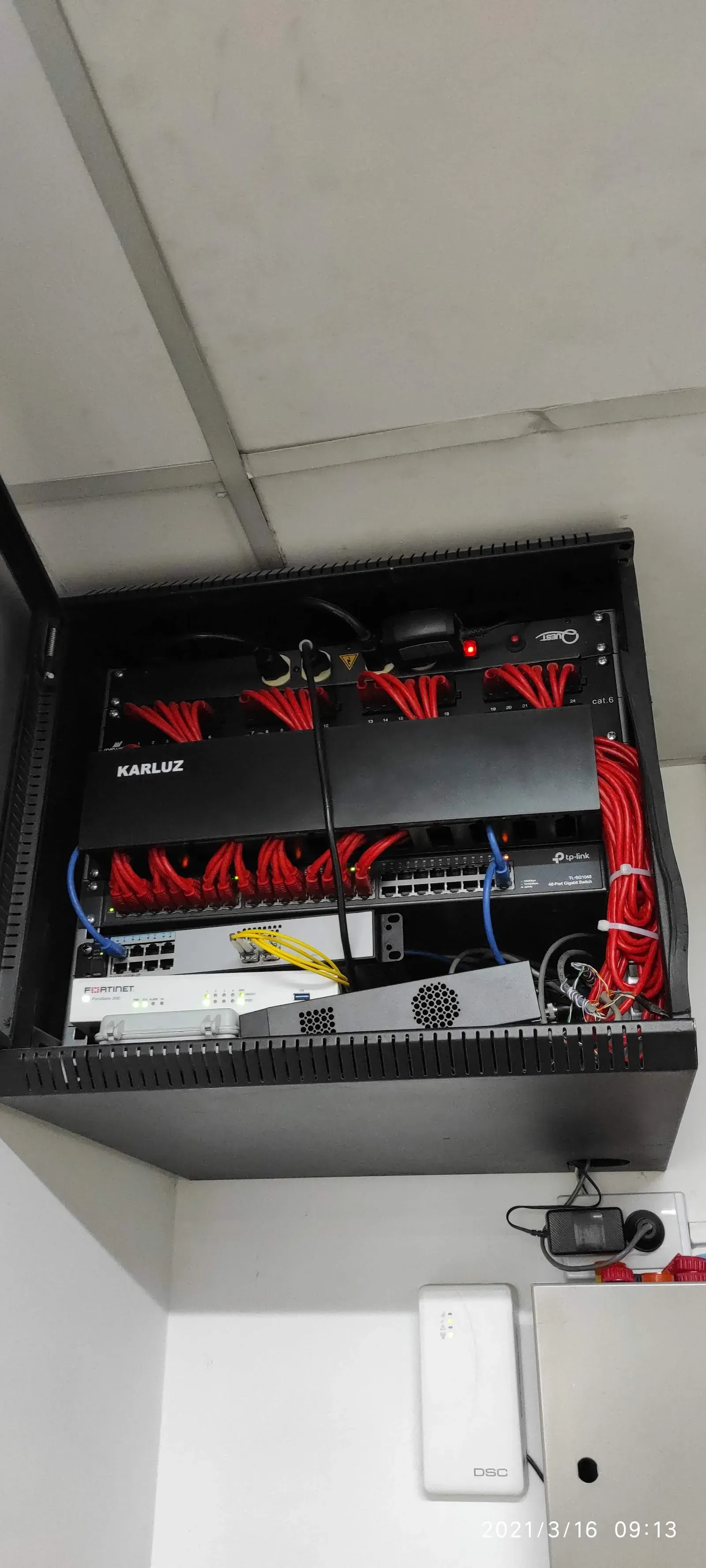 Rack de comunicaciones abierto con patch panel categoría 6 de veinticuatro puertos y cables de parcheo rojos, un switch TP-Link de cuarenta y ocho puertos gigabit, un firewall Fortinet FortiGate y dos cables de fibra óptica amarillos conectados