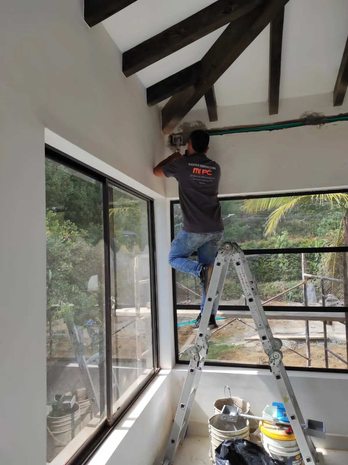 Técnico de MiPC Tecnología, subido en una escalera plegable, instala un punto eléctrico bajo el techo de vigas de madera de una vivienda en construcción