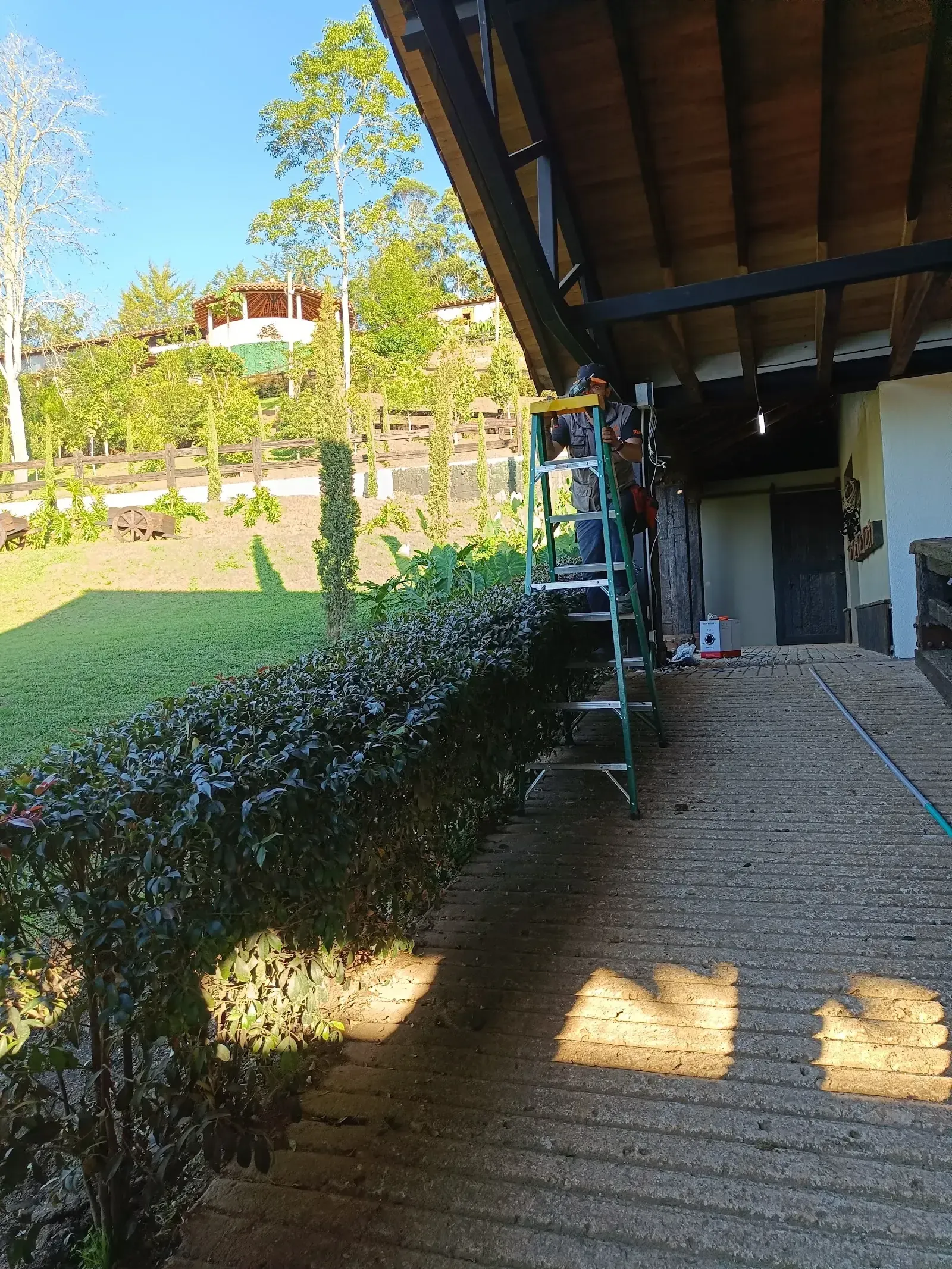 Técnico de MiPC Tecnología, subido en una escalera de fibra de vidrio, instala una cámara bajo el alero de madera de una construcción rural, con el jardín y el paisaje de la propiedad al fondo