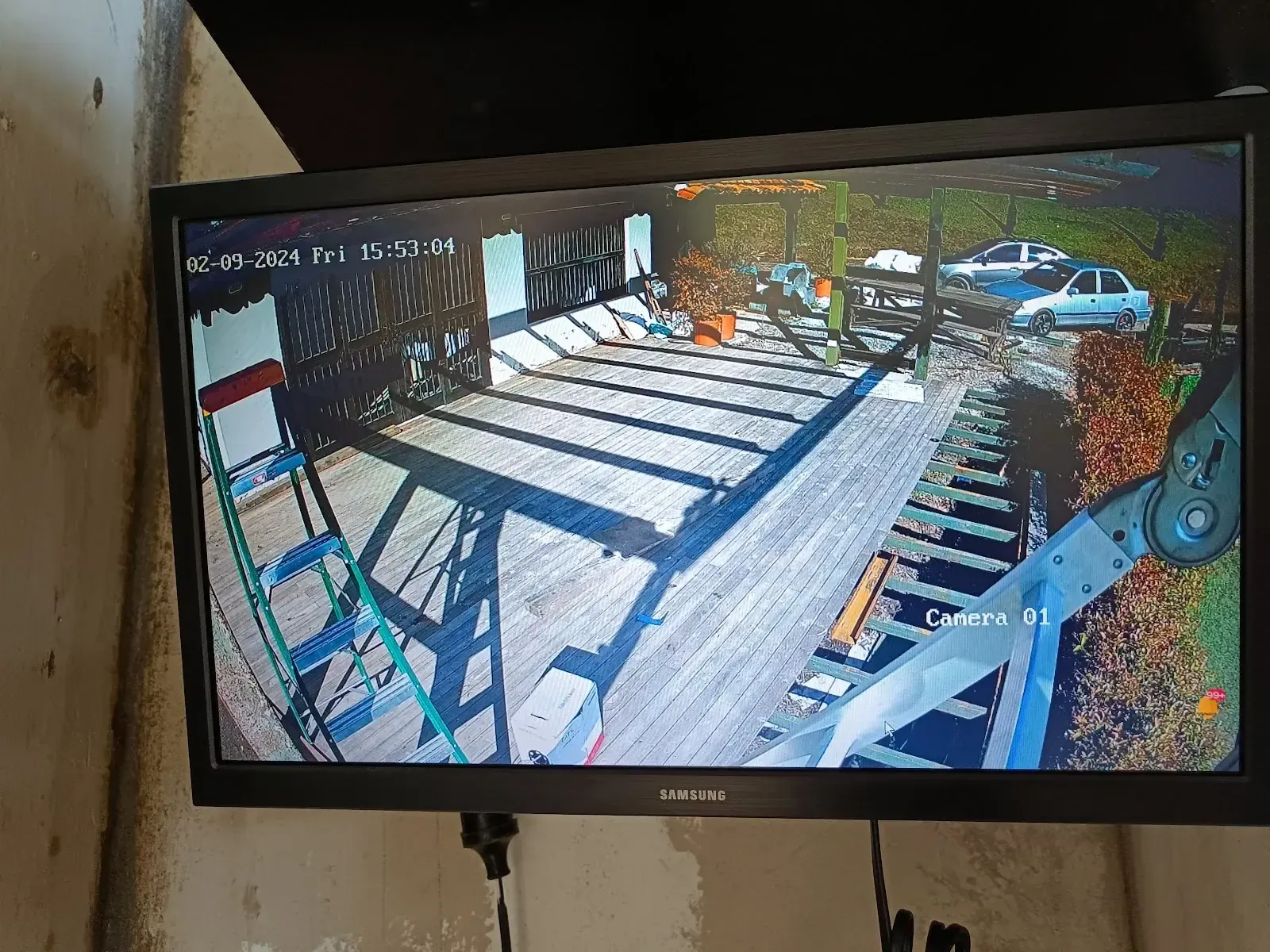 Monitor instalado en la pared mostrando en vivo la imagen de una cámara de seguridad, con la vista del parqueadero y la zona cubierta de la propiedad, y la fecha y el rótulo «Camera 01» sobreimpresos