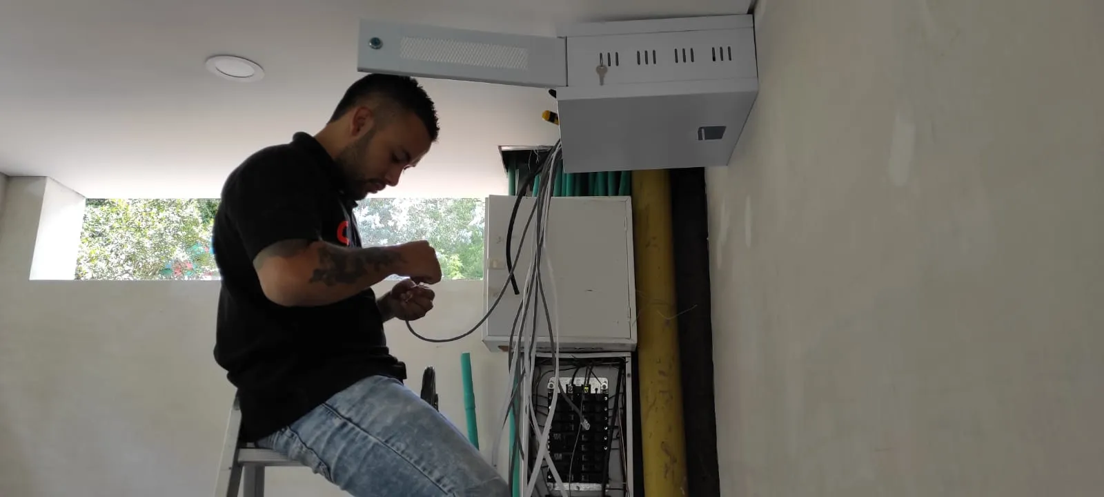 Técnico de MiPC Tecnología, subido en una escalera baja, poncha un cable de red junto a un tablero eléctrico y una caja de paso cerca del techo