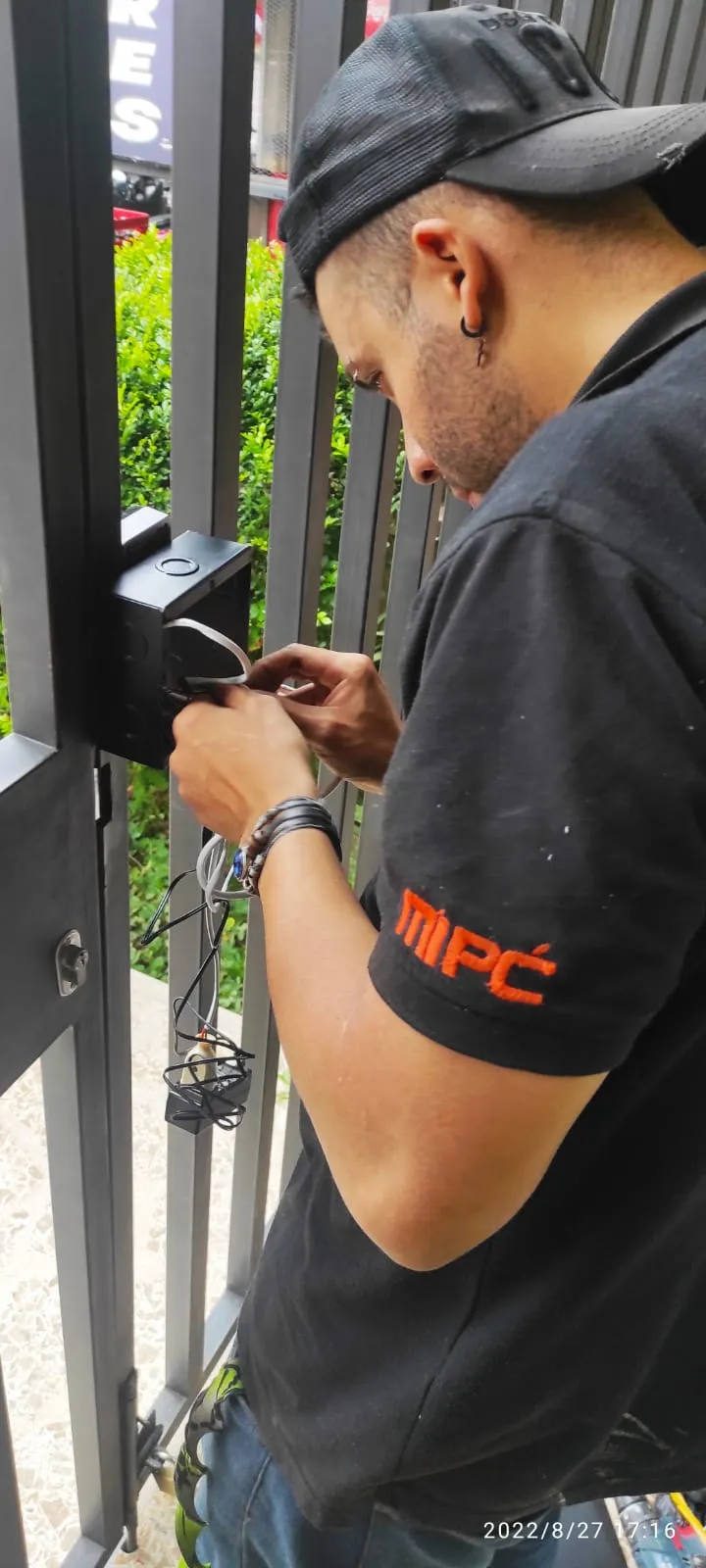 Técnico de MiPC Tecnología con gorra y camiseta de uniforme conecta el cableado de un control de acceso en el marco metálico de un portón