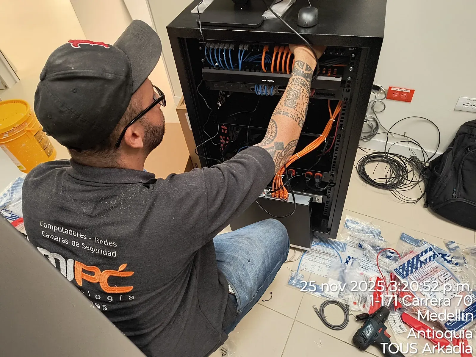 Técnico de MiPC Tecnología, con camiseta del uniforme de la empresa, conecta los cables de parcheo naranjas y azules a un patch panel categoría 6 dentro del rack de la tienda, con un switch Hikvision montado justo debajo