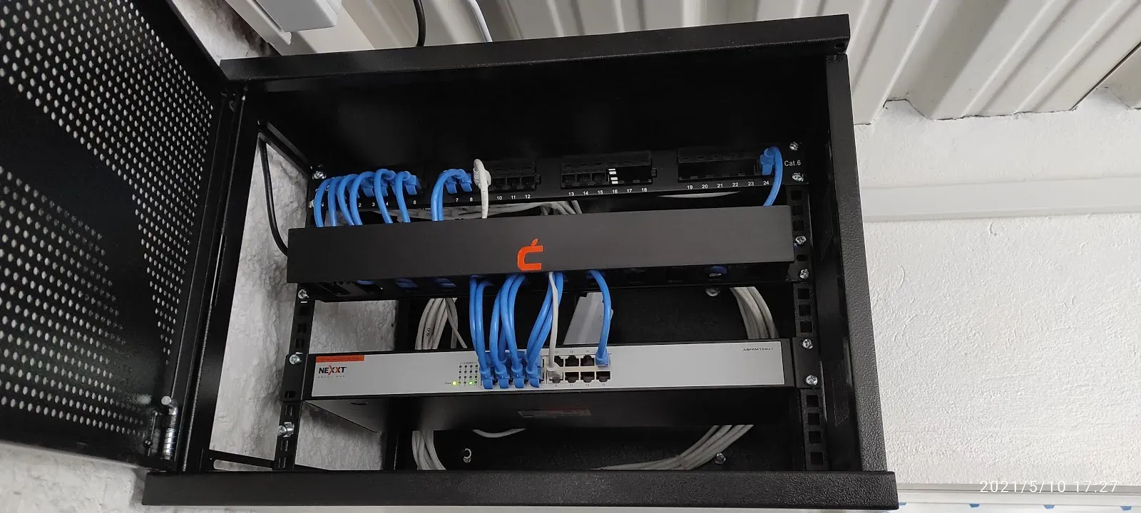 Gabinete de red de pared abierto y terminado, con patch panel categoría 6 de veinticuatro puertos, cables de parcheo azules peinados y un switch Nexxt de ocho puertos montado debajo